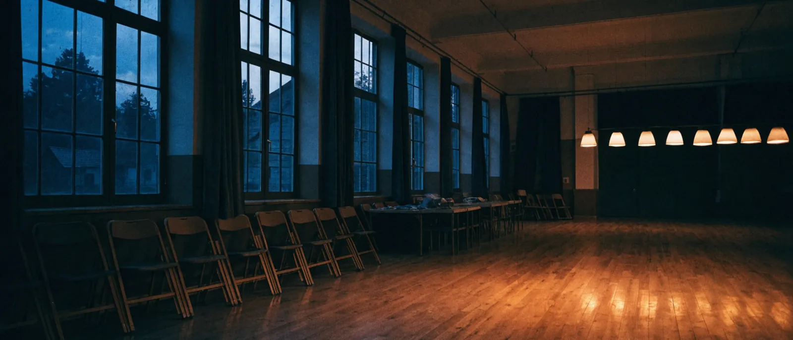 Pusta sala wiejskiej świetlicy o zmierzchu ze złożonymi krzesłami pod ścianą.